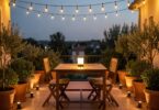 Terrasse illuminée le soir avec guirlandes LED, spots solaires au pied des plantes et lampe nomade sur la table